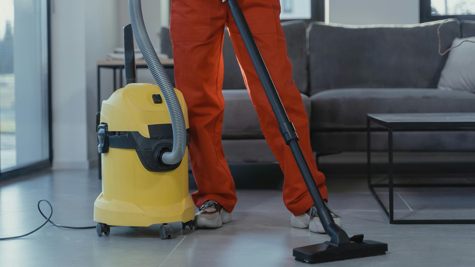 Cleaner in red coveralls vacuuming a smooth indoor floor with a canister vacuum; face outside the frame