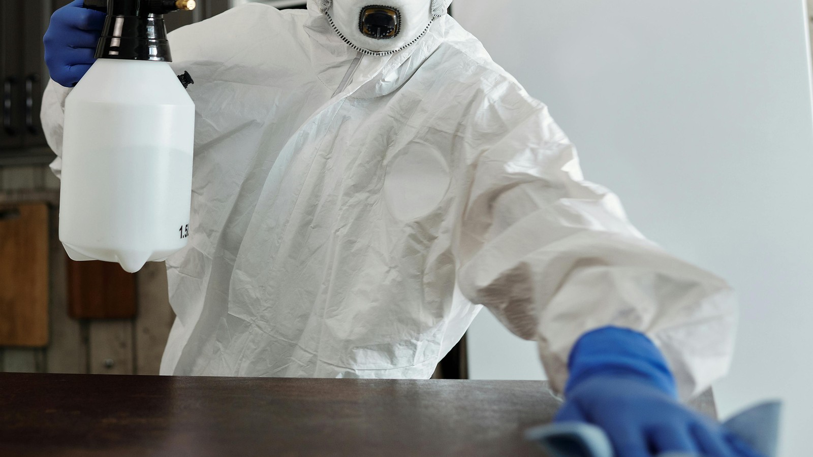 Blue-gloved cleaner in protective coveralls wiping a tabletop with a cloth and spray bottle; face outside the frame