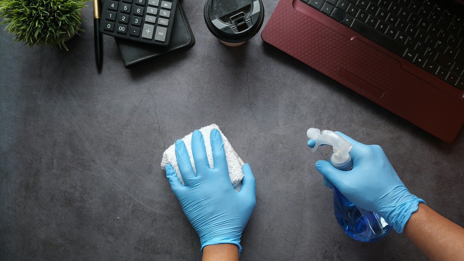 Blue-gloved hands wiping an office desk with a cloth and cleaning spray beside a laptop